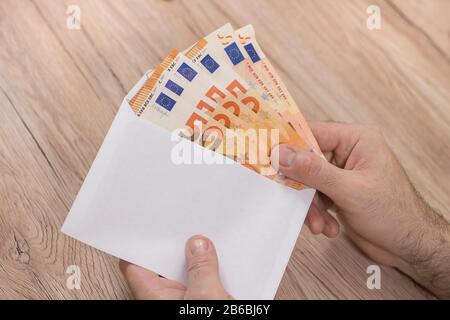 Die Hand eines Mannes hält einen weißen Umschlag und steckt darin Euro-Banknoten mit 50 Euro Stückelung. Stockfoto