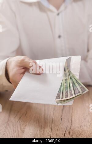 Die Hand eines Mannes hält einen weißen Umschlag, der mit Banknoten der polnischen Währung gefüllt ist, und übergibt ihn über den Tisch. Stockfoto