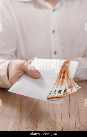 Die Hand des Mannes hält einen weißen Umschlag mit Euro-Banknoten und übergibt ihn über den Tisch. Stockfoto