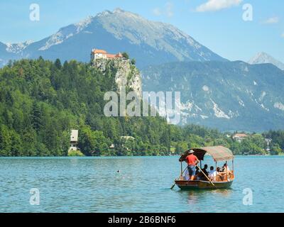 Boot mit Touristen segelt auf dem Bleder See in Slowenien mit Bergen und Schloss im Hintergrund. Stockfoto