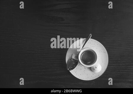 Blasses Schwarzweißbild einer Tasse Espresso. Frisch zubereiteter, starker italienischer Kaffee wird im Restaurant serviert, von oben auf schwarze Holztellstruktur geschossen. Stockfoto