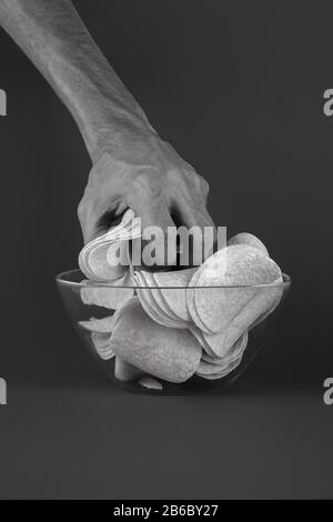 Kartoffelchips aus einer Glasschale holen. Die männliche Hand nimmt eine Handvoll Chips in schwarzem Monochrom-Hintergrund Stockfoto