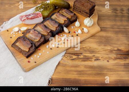 Speck- und Petersilienscheiben auf schwarz geröstetem Brot, Schweinespeck mit Gewürzen und Knoblauch stehen auf einem dunklen Stand. Eingelegte Gurken mit Dill, Knoblauch und Pfirsich Stockfoto