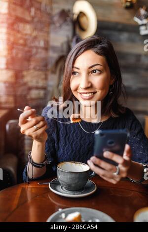 Schöne junge asiatin, die in einem Café sitzt und leckeren Kuchen isst Stockfoto