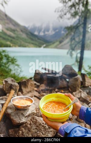 Frau Wanderer, die das Abendessen in einer Schüssel mit heißer Suppe in der Nähe des Bergsees genießen Stockfoto