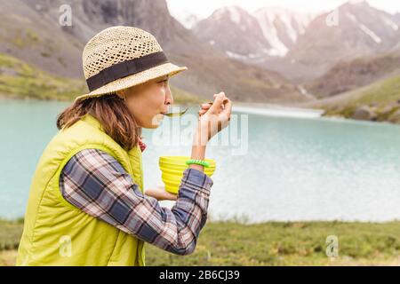 Frau Wanderer, die das Abendessen in einer Schüssel mit heißer Suppe in der Nähe des Bergsees genießen Stockfoto