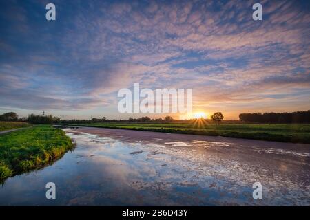 Typische holländische Polderlandschaft mit Reflexion eines mehrfarbigen Sonnenaufgangs am Wasser in einem breiten Graben. Stockfoto