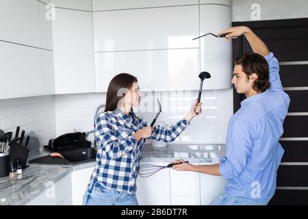 Lustige Paare, die vorgeben, mit Utensilien zu kämpfen, während sie zu Hause kochen, haben Mann und Frau Spaß dabei, spielerisch die küchen-stru zu halten Stockfoto