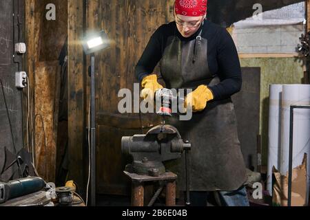 Die Handwerkerin handhabt ein Metallkunstwerk, das in einem Schraubstock mit einem Winkelschleifer in einer Werkstatt eingespannt ist Stockfoto