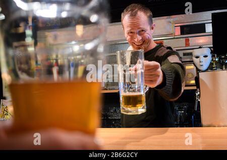 Rothaariger fast irischer Kerl, der hinter der Bar barzt und mit etwas Bier jubelt und mit Smiley-Gesicht auf Sie und Ihren Jug schaut Stockfoto