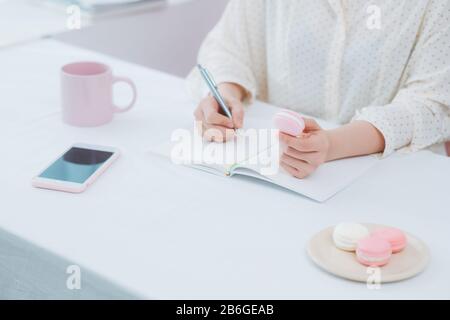 Nahaufnahme von Frauen oder Studenten, die mit Bleistift, Becher, Makronen und Telefon in Notizbuch schreiben Stockfoto