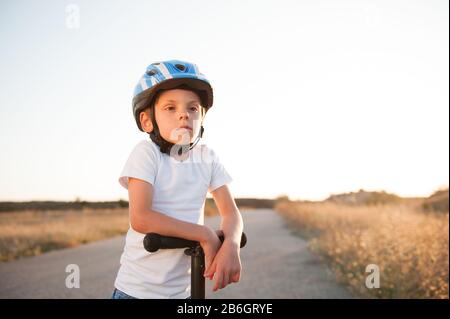 Portrait des kleinen Jungen in Sporthelm und Roller, der auf der leeren Straße zum Sonnenuntergang in der Wüste steht Stockfoto