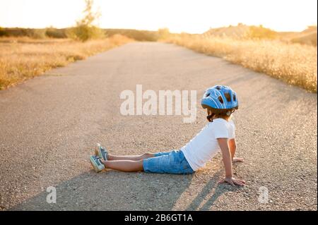 Aktives kleines Kind mit Sport Outfit Helm und Turnschuhe sitzen auf leerer Asphaltstraße bei sommerlichem warmen Sonnenuntergang Stockfoto