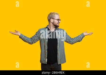 Bärtiger kaukasischer Mann mit blondem Haar vergleicht zwei Dinge von seinen Handflächen, während er auf einer gelben Wand posiert Stockfoto