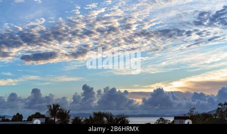 Sonnenaufgang als die Dämmerung zwischen den Wolken hinter den Bergen und über dem Ozean bricht, schöne Wolken gegen einen weichen blauen Himmel Stockfoto