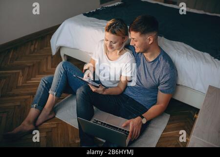 Obere Ansicht des kaukasischen Paares, das auf dem Boden sitzt und moderne Geräte verwendet, während sie lächeln und Freude haben Stockfoto