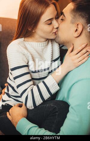Liebliches rothaariges Mädchen mit Freckles umarmt ihren Liebhaber und küsst, während es von ihm auf einem Sofa umarmt wird Stockfoto