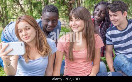 Gruppe gemischter Rennen Teenager, die mit Smartphone auf dem Gras pfiffen, um Selfies zu machen. Stockfoto