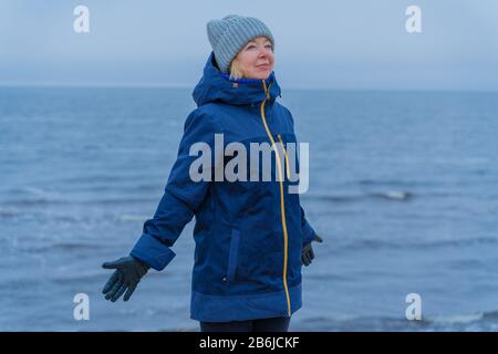 Kalter Wintertag. Porträt einer Frau mittleren Alters mit ausgestreckten Händen auf blauem Meeresgrund Stockfoto