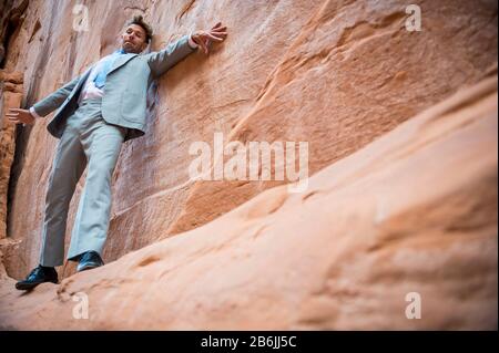 Der nervöse Geschäftsmann klammert sich an ein Felswand und balanciert auf einem schmalen Felsvorsprung in einem roten Felsschlucht Stockfoto