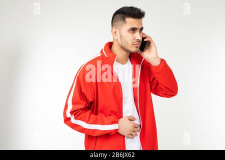Brünette stylische Mann in einer roten Sportjacke spricht auf einem Smartphone auf einem weißen leeren Hintergrund Stockfoto