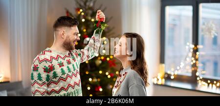 Weihnachten, Menschen und Urlaubstradition Konzept - Porträt des glücklichen Paares in hässlichen Pullovern mit Misteln über den Heimathintergrund Stockfoto