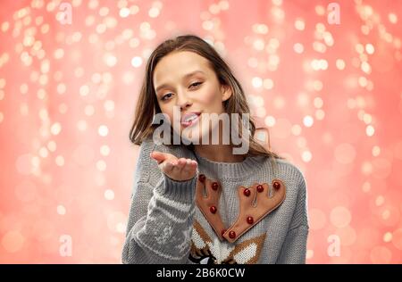 Weihnachten, Menschen und Urlaub Konzept - fröhliche junge Frau mit hässlichem Pullover mit Rentiermuster, die einen Luftkuss über festliche Lichter auf pinkfarbener Kor sendet Stockfoto