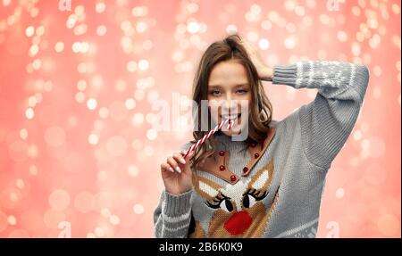 Weihnachten, Menschen und Urlaub Konzept - fröhliche junge Frau mit hässlichem Pullover mit Rentiermuster, die Süßigkeiten über festlichen Lichtern auf pinkfarbenem Co beißt Stockfoto