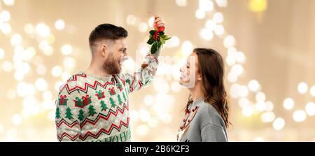 Weihnachten, Menschen und Urlaubstradition Konzept - Porträt des glücklichen Paares in hässlichen Pullovern mit Misteln über festlichem Lichterhintergrund Stockfoto