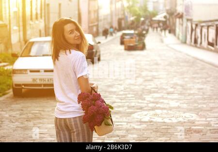 Stilvolle Frau mit Stroh Beutel mit einem lebendigen Haufen lila Blumen und auf der Straße. Moody sonnigen Portrait. Stockfoto