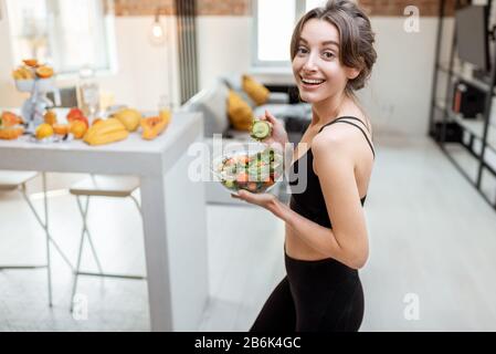 Porträt einer fröhlichen athletischen Frau, die während einer Pause zu Hause gesunden Salat isst. Konzept des Gewichtsverlustes, des Sports und des gesunden Essens Stockfoto