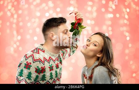 Weihnachten, Menschen und Urlaub Traditionen Konzept - Porträt des glücklichen Paares in hässlichen Pullover, die unter Misteln über festliche Lichter auf rosa Koralle küssen Stockfoto