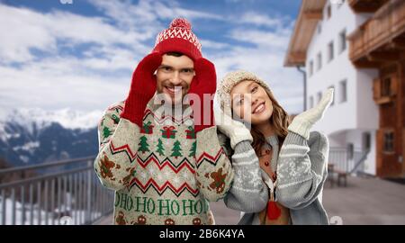 Weihnachten, Winterkleidung und Weihnachtskonzept - glückliches Paar in hässlichen Pullovern, Strickmützen und Fäusten über dem Skigebiet in den österreichischen alpen Berge BA Stockfoto