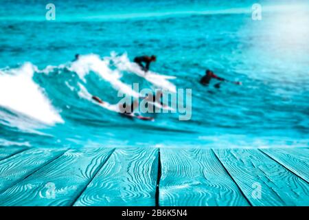 Surfer auf Wellen im türkisfarbenen Ozean. Blauer Holztisch mit Platz für Ihre Dekoration und Werbemittel. Fröhliche aktive sonnige Sommerzeit Stockfoto