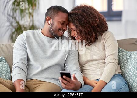 Technologie-, Musik- und Personkonzept - glückliches afroamerikanisches Paar mit Smartphone und Ohrhörer zu Hause Stockfoto