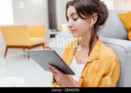 Junge und fröhliche Frau, die mit kabellosen Kopfhörern auf einem digitalen Tablet arbeitet, während sie zuhause entspannt sitzt. Konzept der Freizeit-, freiberuflichen und mobilen Arbeit Stockfoto