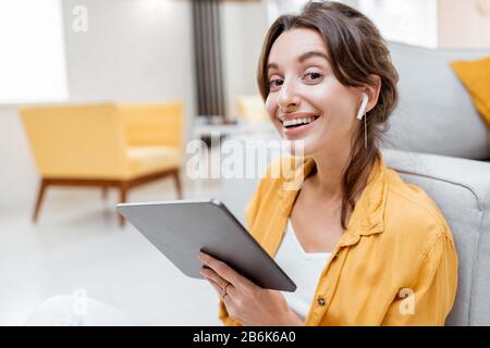 Junge und fröhliche Frau, die mit kabellosen Kopfhörern auf einem digitalen Tablet arbeitet, während sie zuhause entspannt sitzt. Konzept der Freizeit-, freiberuflichen und mobilen Arbeit Stockfoto