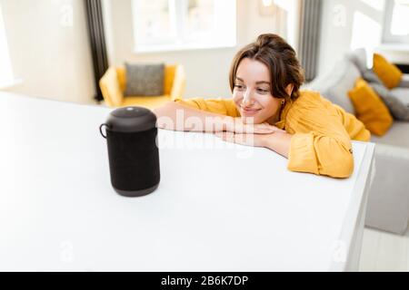 Fröhliche junge Frau steuert intelligente Heimgeräte mit Sprachbefehlen und cleverem Lautsprecher zu Hause. Konzept eines intelligenten Heimnetzhauses und Remote-Verwaltung drahtloser Geräte Stockfoto