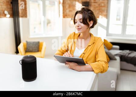 Fröhliche junge Frau steuert intelligente Heimgeräte mit Sprachbefehlen und cleverem Lautsprecher zu Hause. Konzept eines intelligenten Heimnetzhauses und Remote-Verwaltung drahtloser Geräte Stockfoto