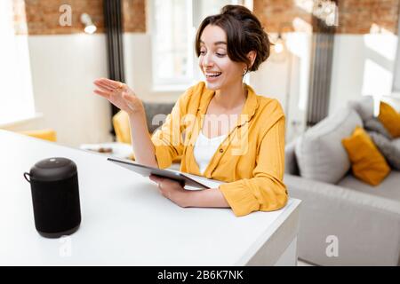 Fröhliche junge Frau steuert intelligente Heimgeräte mit Sprachbefehlen und cleverem Lautsprecher zu Hause. Konzept eines intelligenten Heimnetzhauses und Remote-Verwaltung drahtloser Geräte Stockfoto