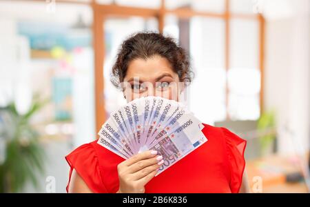 Konzept "Menschen und Finanzen" - glückliche Frau im roten Kleid, die ihr Gesicht hinter Euro-Geldscheinen über Bürohintergrund versteckt Stockfoto