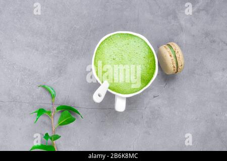 Französische Mandelkuchen-Makarons, grünes Blatt, Matcha-Tee in weißer Tasse auf Betongrund Stockfoto
