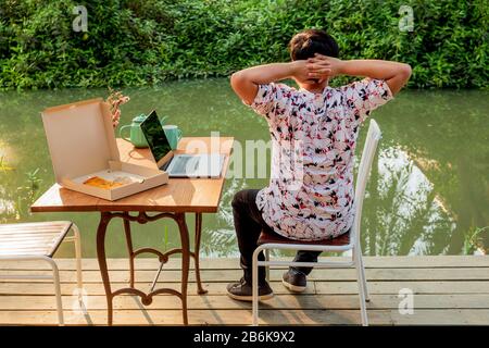 Pizaa auf dem Tisch mit jungen Mann-Händen hinter dem Kopf, nachdem er an einem Laptop gearbeitet hatte. Stockfoto