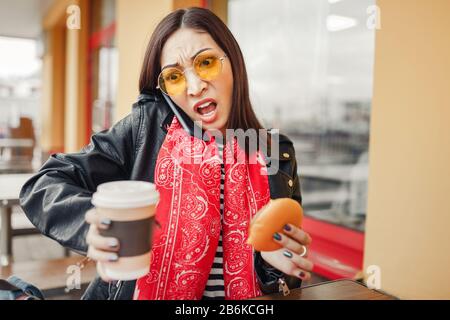 Überraschte beschäftigte Frau, die während eines Abendessens in einem Fast-Food-Restaurant telefoniert Stockfoto