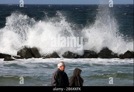 Wustrow, Deutschland. März 2020. Spaziergänger sind auf der Halbinsel Fischland an der turbulenten Ostsee, an einem Wellenbrecher spritzt das Wasser auf. Sonne und Squalls bis zu Force Eight sorgen für eine beeindruckende Kulisse. Credit: Bernd Wüstneck / dpa-Zentralbild / dpa / Alamy Live News Stockfoto