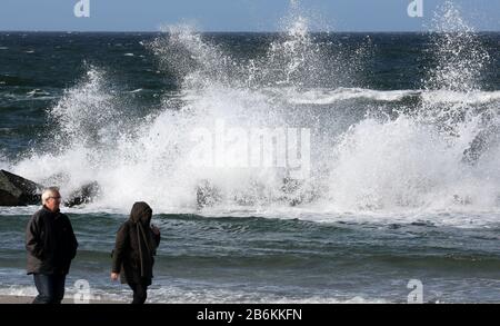 Wustrow, Deutschland. März 2020. Spaziergänger sind auf der Halbinsel Fischland an der turbulenten Ostsee, an einem Wellenbrecher spritzt das Wasser auf. Sonne und Squalls bis zu Force Eight sorgen für eine beeindruckende Kulisse. Credit: Bernd Wüstneck / dpa-Zentralbild / dpa / Alamy Live News Stockfoto