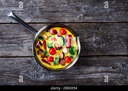 Gebackene Omelette oder Frittata mit Würstchen, Brokkoli, Kirschtomaten, geschmolzenem Käse im Skillet auf einem Holztisch im Landhausstil, horizontale Sicht von oben, fl Stockfoto