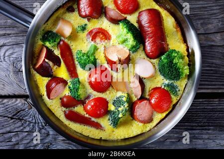 Gebackenes Omelett oder Frittata mit Würstchen, Brokkoli, Kirschtomaten, geschmolzenem Käse im Skillet auf einem Holztisch im Landhausstil, horizontale Ansicht von oben, Stockfoto