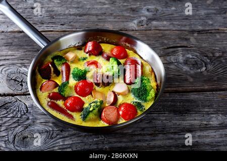 Gebackene Omelette oder Frittata mit Würstchen, Brokkoli, Kirschtomaten, geschmolzenem Käse in einem Skillet auf einem Holztisch im Landhausstil, horizontale Ansicht von oben Stockfoto