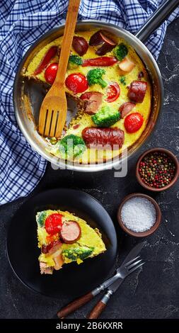 Gebackene Omelette oder Frittata mit Würstchen, Brokkoli, Kirschtomaten, geschmolzenem Käse in einem Skillet auf einem Betontisch und einer Portion auf einem Teller, senkrecht Stockfoto